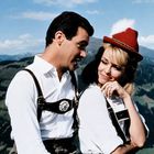 Ein Mann und eine Frau in Tracht, beide lächeln sich vor einer alpinen Landschaft an.