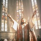 Ein Priester in einem prächtigen Gewand erhebt die Arme in einer Kirche mit farbenfrohen Buntglasfenstern.