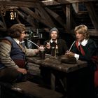 Drei Personen sitzen in rustikaler Umgebung, rauchen lange Pfeifen. Eine Kerze brennt auf einem Holztisch.