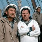 Auf Matrosenmützen steht "Михайлов". Zwei Männer in Seemannskleidung auf einem Schiff, schauen ernst in die Ferne.