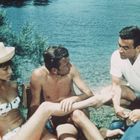 Es sind keine Texte vorhanden. Drei Personen am Ufer, eine Frau im Bikini und zwei Männer in Badekleidung in Gespräch vertieft.