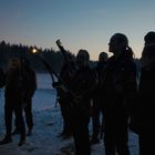 Menschen halten Fackeln in einer verschneiten Landschaft bei Dämmerung, umgeben von einem Wald im Hintergrund.