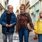 Drei Personen gehen eine alte Gasse entlang. Eine Frau hält eine kleine Trommel. Ein Mädchen trägt eine gelbe Jacke.