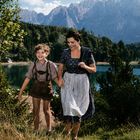 Keine sichtbaren Texte. Eine Mutter und ihr Sohn spazieren lachend, in traditioneller Kleidung, vor einer Berglandschaft mit See.