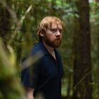 Keine erkennbaren Texte. Ein rothaariger Mann mit Bart steht in einem dichten, grünen Wald und schaut ernst.