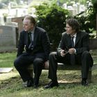 Zwei Männer in dunklen Anzügen sitzen auf einer Bank auf einem Friedhof, umgeben von Grabsteinen mit den Namen "KUDDEBACK" und "HAND".