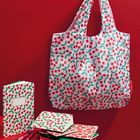 An einer roten Wand hängt eine Tasche mit Kirschmuster. Darunter Notizbücher mit demselben Muster auf einem Tisch.