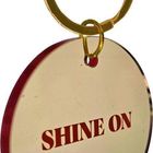 Auf einem runden Anhänger steht in roten Großbuchstaben "SHINE ON". Der Anhänger ist an einem goldfarbenen Schlüsselring befestigt.
