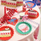 Papiertüten und Teller in Form von Tortenstücken, Blumenmotiv und Schleifen in Rosa-, Rot- und Blautönen.