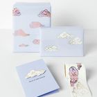 „HEAD IN THE CLOUDS“ auf einer hellblauen Karte mit Wolkenmotiv; Geschenkverpackungen und Sticker in Pastelltönen.