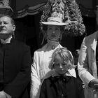 Vier Personen in festlicher Kleidung stehen nebeneinander, darunter ein Kind. Dekorativer Hintergrund mit Pflanzen.