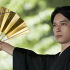 Keine Texte. Ein Mann in traditioneller Kleidung hält einen goldenen Fächer, der Hintergrund ist verschwommen.
