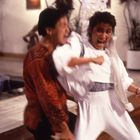 Zwei Personen in einem energischen Kampf, die Frau trägt Weiß, der Mann ein rotes Hemd, Innenraum mit Pflanzen.