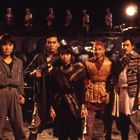 Fünf Personen in Abenteurerkleidung schauen aufmerksam, im Hintergrund Soldaten in Uniform auf einer Mauer.