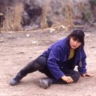 Frau in blauer Jacke und Stiefeln sitzt am Boden in einer staubigen Umgebung. Gras und Felsen im Hintergrund.