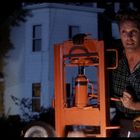 Ein Mann in kariertem Hemd steht draußen und hält einen orangefarbenen Wagenheber. Die Umgebung wirkt dunkel und beleuchtet.