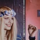 Eine Frau mit langen, blonden Haaren und Blumenkranz lächelt, während sie ihre Hand leicht hebt. Hintergrund in Rot- und Blautönen.