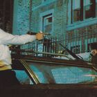 Zwei Personen mit Waffen in einem städtischen Hinterhof, nahe einem Auto mit Ziegelgebäude im Hintergrund, Nachtstimmung.