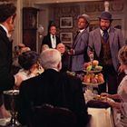 Es gibt keinen erkennbaren Text. Zwei Männer im Vordergrund halten Pistolen, während andere Personen an einem gedeckten Tisch sitzen.