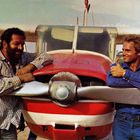 Zwei Männer lehnen lächelnd an einem kleinen roten Flugzeug, einer zeigt Daumen hoch.