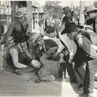 "Bank of Texas" steht auf einem Schaufenster. Mehrere Männer in Cowboykleidung kümmern sich um jemand, der am Boden liegt.
