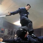 Zwei Männer kämpfen; einer tritt auf den anderen, während er einen Stock hält. Die Szene wirkt angespannt und dynamisch.