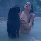 Zwei Personen umarmen sich im nebligen Wasser, umgeben von schwimmenden Blütenblättern und felsiger Landschaft im Hintergrund.