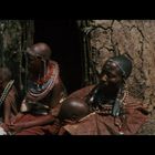 Keine Texte vorhanden. Vier Personen in traditioneller Kleidung sitzen vor einer Hütte aus Lehm.