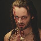 Ein Mann mit blutverschmiertem Gesicht und langem Haar, trägt eine Kette mit einem auffälligen Anhänger.