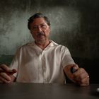 Ein Mann in einem weißen Hemd sitzt an einem Tisch und hält eine Zigarre. Der Hintergrund ist düster und strukturiert.