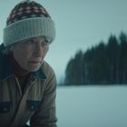 Eine ältere Person in Winterkleidung und Mütze, vor einem verschneiten Hintergrund und Wald.