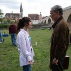 Eine junge Frau und ein älterer Mann stehen sich auf einer Wiese gegenüber. Im Hintergrund ist eine alte Brücke und eine Kirche.