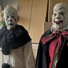 Zwei Figuren in Clownkostümen. Links trägt ein weißes Outfit mit schwarzem Kragen, rechts ein dunkles Cape mit rotem Schal.