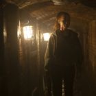 Kein Text zu sehen. Eine Person mit hochgestecktem Haar geht durch einen dunklen, ziegelsteinernen Tunnel mit Laternenlicht.