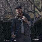 Ein Mann steht draußen, hält eine Schaufel und trägt eine gepolsterte Jacke und Handschuhe. Im Hintergrund sind kahle Bäume.