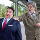 Auf einem Bahnsteig steht eine Frau in Uniform mit Namensschild "Kulick", während ein Mann ihren Rücken antippt.