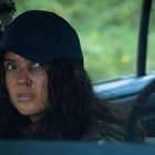 Eine Frau mit blutigen Kratzern im Gesicht sitzt in einem Auto, trägt eine dunkle Kappe und schaut nachdenklich zur Seite.