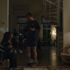 Ein Mann steht in Shorts mit gesenkter Hose; eine Frau sitzt und schaut ihn an; dahinter eine stilvolle Wohnung.