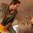 Ein Mann mit Bart hält einen gelben Feuerlöscher in aggressiver Haltung über einen anderen Mann, der blutet und sitzt.