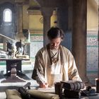 Ein Mann in traditioneller Kleidung schreibt mit einer Feder. Er steht in einem Raum mit verzierten Wänden und alten Büchern.