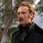 Ein Mann mit Schnurrbart und viktorianischer Kleidung steht draußen vor Bäumen und blickt nachdenklich.