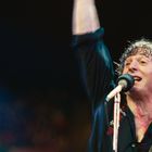 Ein Sänger hält ein Mikrofon und freut sich, während er seine Hand in die Luft hebt, bunte Lichter im Hintergrund.