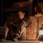 Ein Mann sitzt in einem dunklen Raum, liest ein Buch unter einer Laterne, umgeben von Kartons und Werkzeugen.