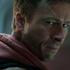 Ein Mann mit ernstem Gesichtsausdruck blickt über die Schulter, trägt einen roten Kapuzenpullover.