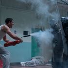 Ein Mann im weißen Tanktop benutzt einen Feuerlöscher gegen eine Reihe von Polizisten mit Schutzschildern.