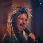 Ein Mann mit wildem, blonden Haar singt leidenschaftlich in ein Mikrofon, umgeben von bunten Lichtern.
