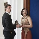 Ein Mann und eine Frau in eleganter Kleidung schauen überrascht, während sie ein silbernes Teeservice halten.