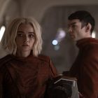 Zwei Personen in futuristischen, rotbraunen Anzügen in einer Raumstation. Eine Frau mit blondem Haar schaut ernst.