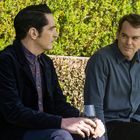 Zwei Männer sitzen nebeneinander, einer hält ein Weinglas. Sie schauen sich an, im Hintergrund sind grüne Büsche.