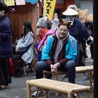 Schilder mit Preisen in Yen; Menschen in Kostümen; Mann mit Fuchsohren und bemaltem Gesicht sitzt auf Bambusbank.